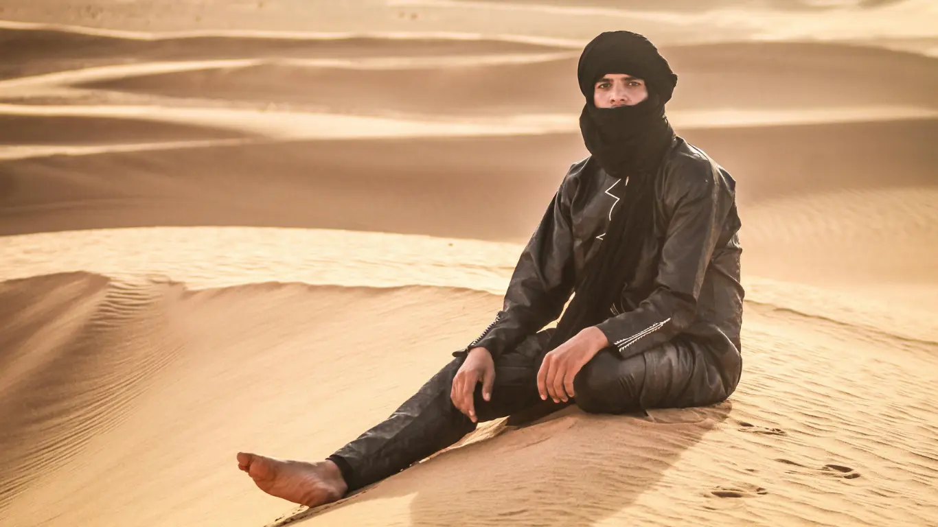 Man dressed in traditional Berber attire sitting on a sand dune in the Sahara Desert during a 3 Days From Marrakech To Merzouga