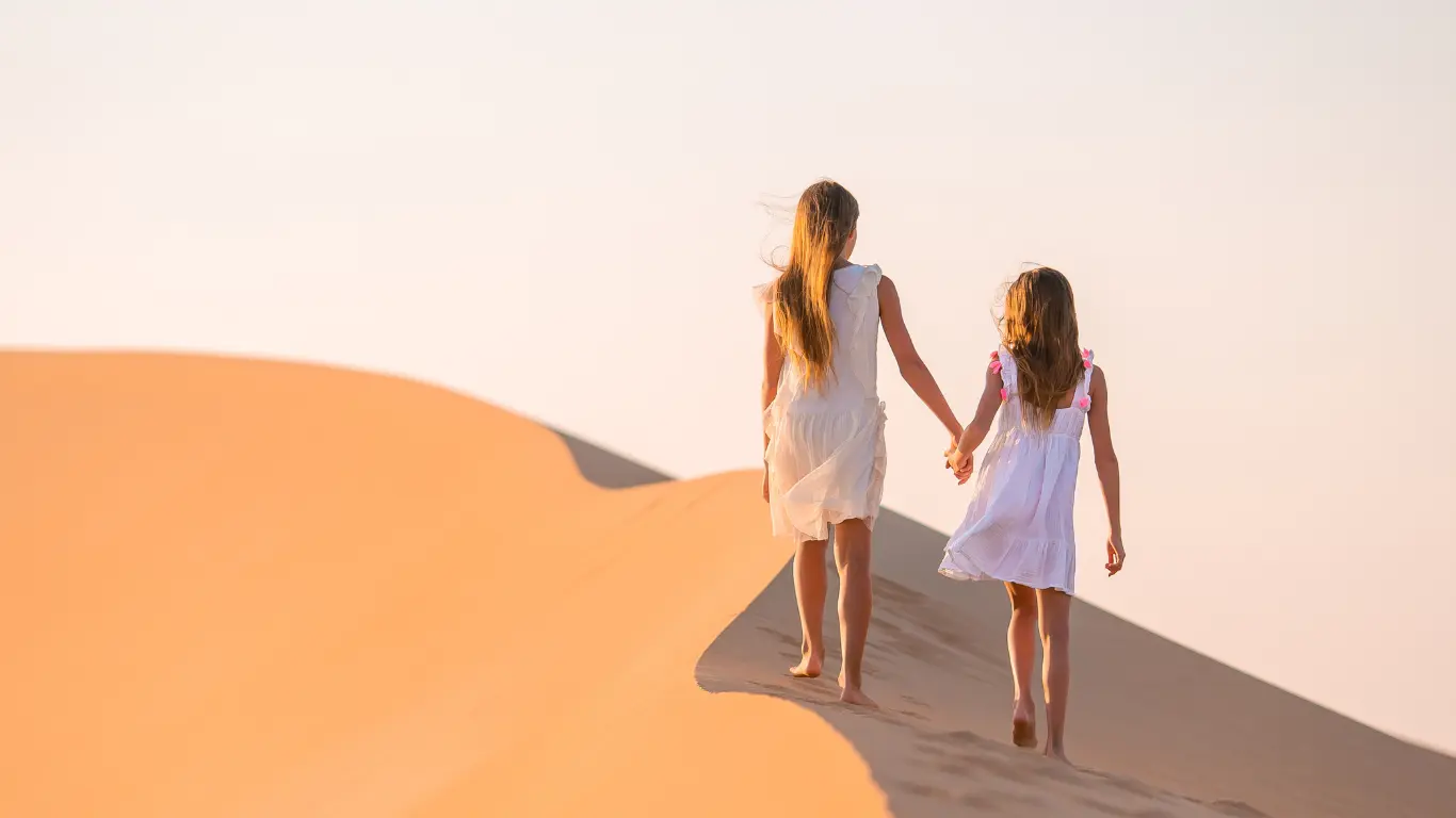 Two girls walking hand-in-hand on the golden sand dunes of Merzouga during a sunset on a desert tour from Marrakech.