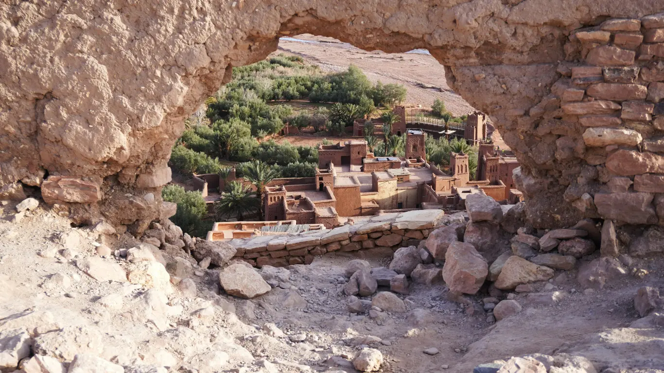 Aït Benhaddou seen through a stone archway, showcasing ancient kasbah architecture and lush greenery during a Marrakech to Merzouga tour.