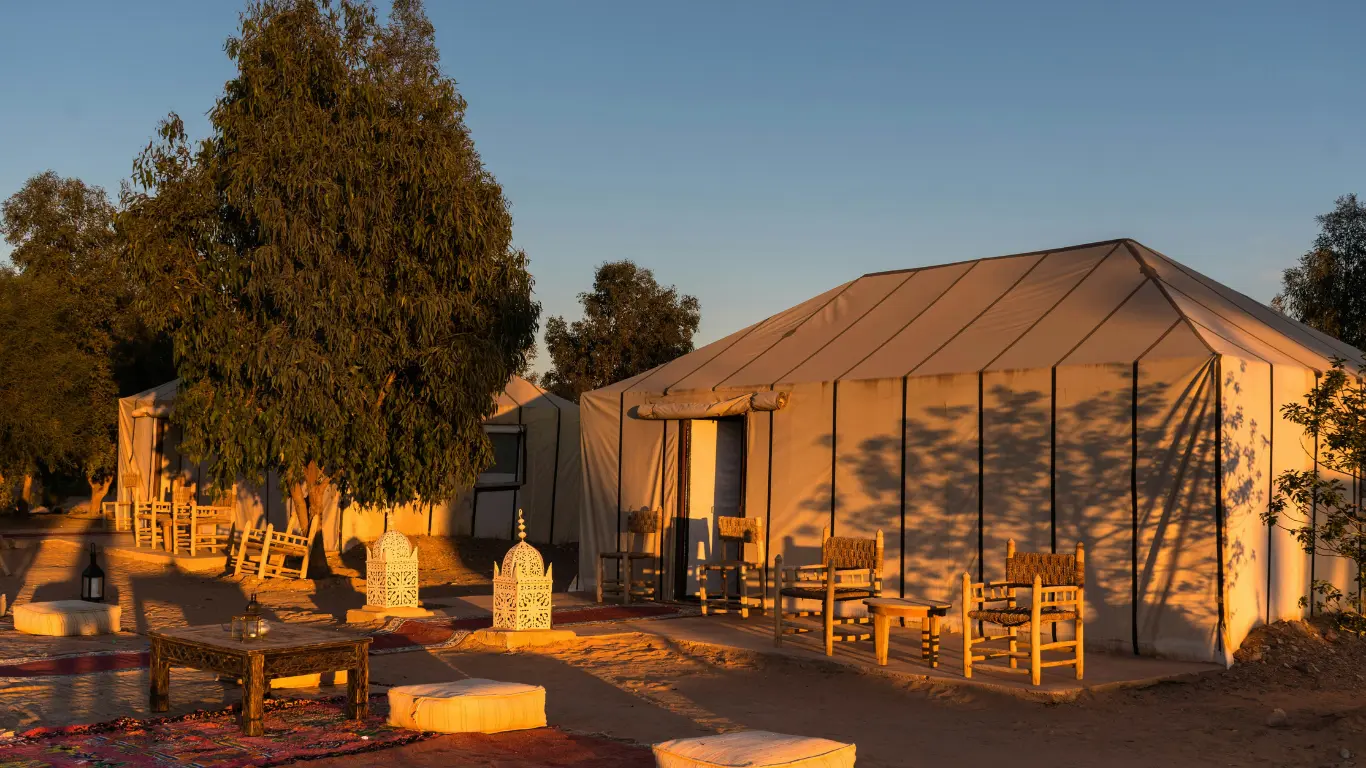 Desert luxury camp at golden hour with Berber tents, wooden chairs, lanterns, and rugs, part of a 4 DAYS TOUR FROM MARRAKECH TO FES.