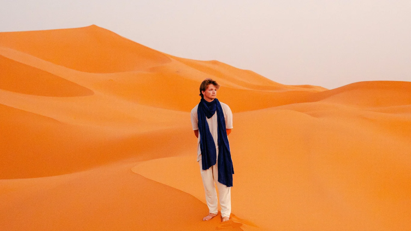 Man in traditional desert attire standing barefoot on orange sand dunes in the Merzouga Sahara, during a 3-day Morocco desert tour from Marrakech to Fes.