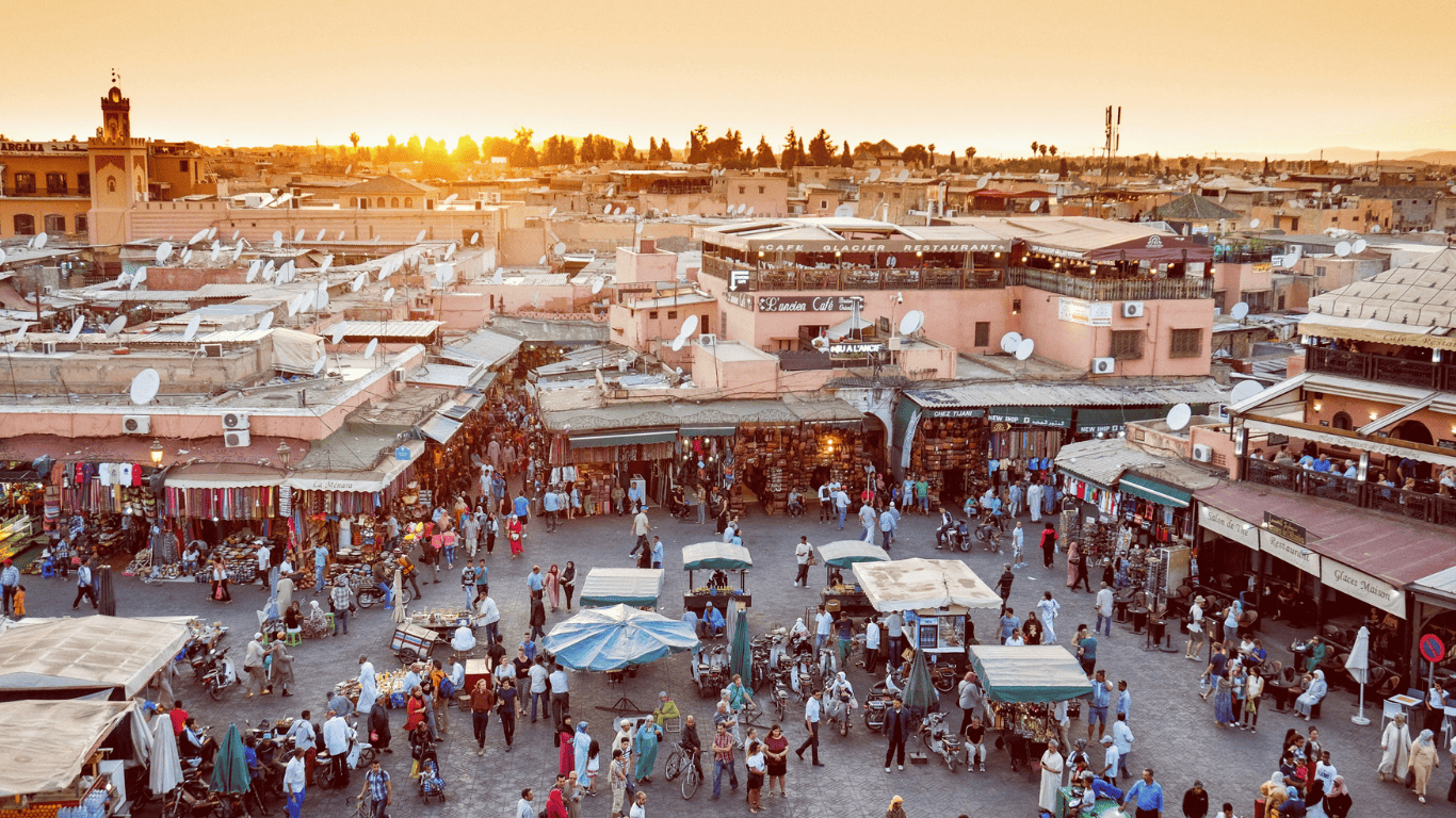 Marrakech Desert Tours Bustling marketplace in Marrakech at sunset, with people shopping, food stalls, and traditional buildings.