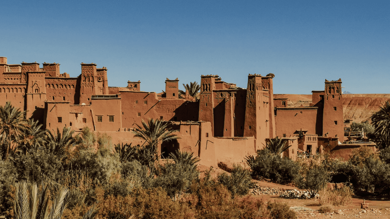5 day desert tour from Marrakech – Historic Ait Ben Haddou kasbah surrounded by palm trees under a clear blue sky.