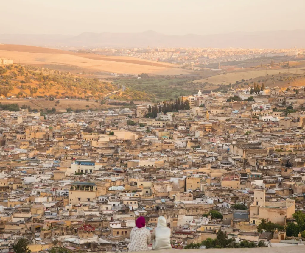 Sunset Over Fes – A Cultural Highlight of Your 3 Day Morocco Tour Panoramic view of Fes Medina at sunset, with two women in traditional dress admiring the city a serene moment on a 3 day Morocco tour.
