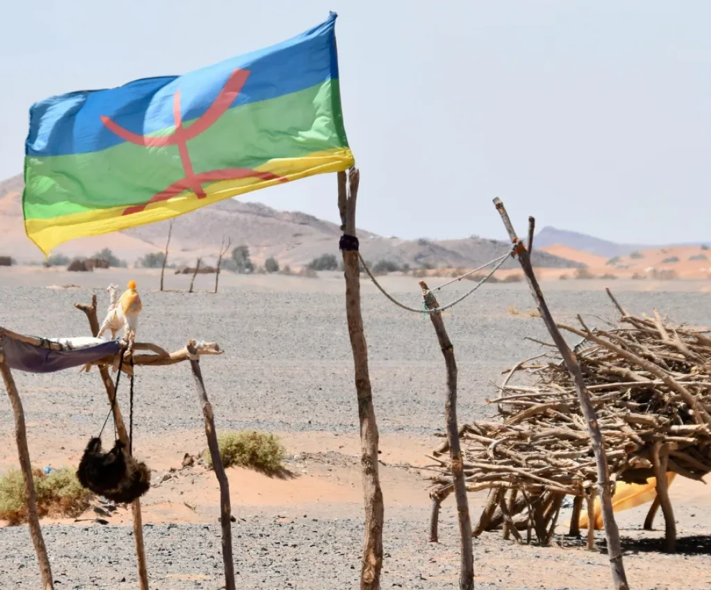 Cultural Moments on a 3 Days Tour from Marrakech to Fes A Berber flag flying in the desert wind near a traditional nomadic shelter made of wood and cloth, with the Atlas Mountains and Sahara dunes in the background.