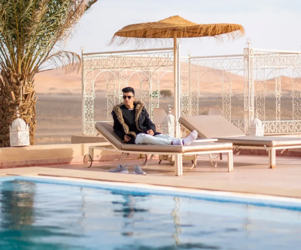 Luxury Desert Escape – 4 Days in Morocco A man relaxing on a poolside lounge chair in the Moroccan desert, surrounded by palm trees and traditional decor, under a straw parasol during a sunny day.