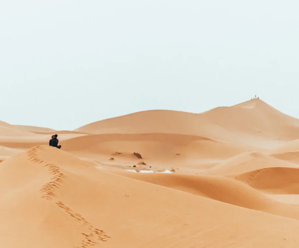 A lone traveler sits atop a vast sand dune in the Moroccan Sahara, gazing out over the undulating golden desert landscape.